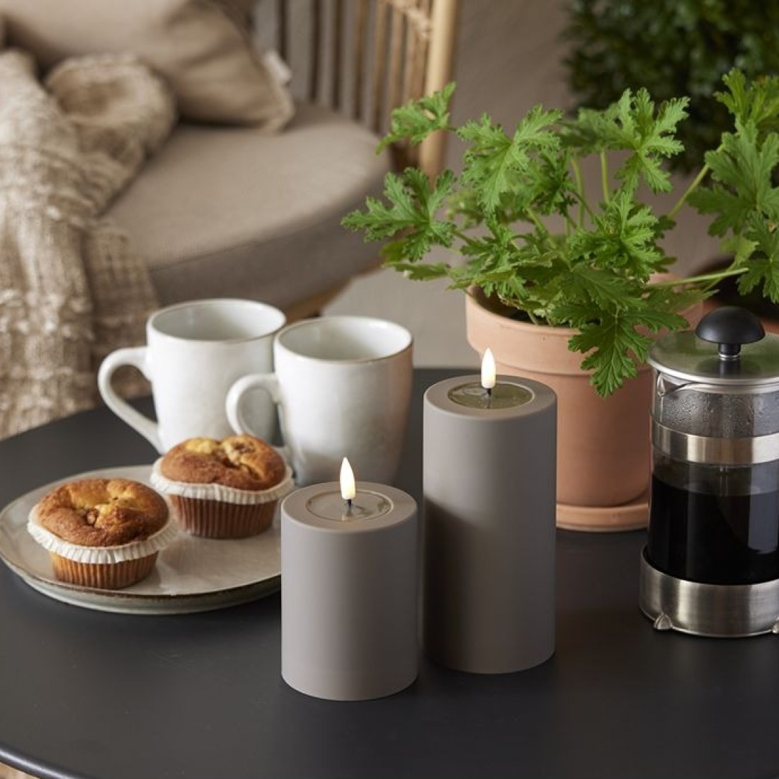 Szare świece LED walcowe obok filiżanek, ciastek i kawiarki na stoliku w otoczeniu zielonych roślin.