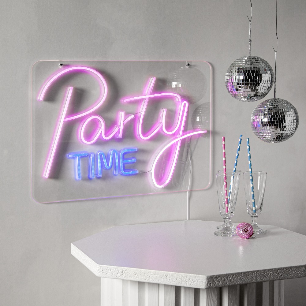 Neon LED Party Time świecący na różowo i niebiesko, zawieszony na szarej ścianie. Pod nim biały stolik, wiszące kule disco, dwa kieliszki ze słomkami i mała różowa kula.