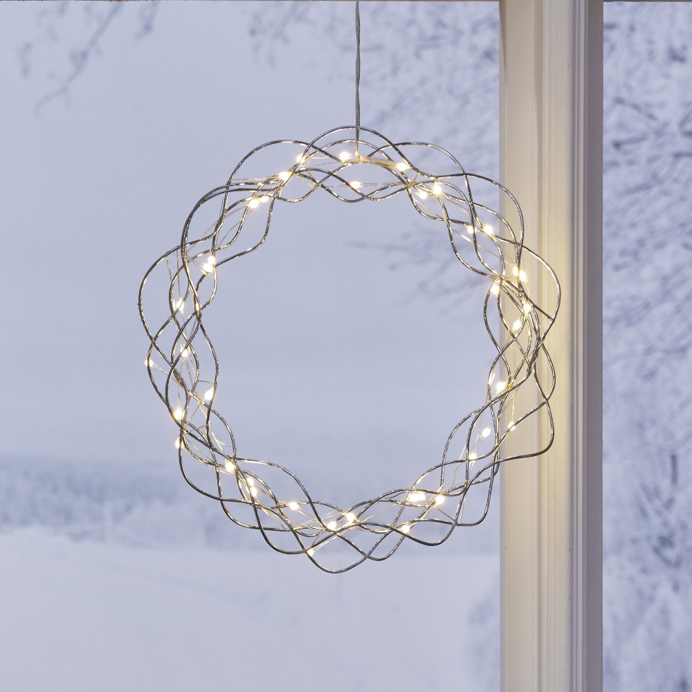 Srebrny wianek LED wykonany z cienkich, przeplatających się drucików, podświetlony ciepłymi punktami światła i zawieszony w oknie z widokiem na ośnieżony krajobraz.