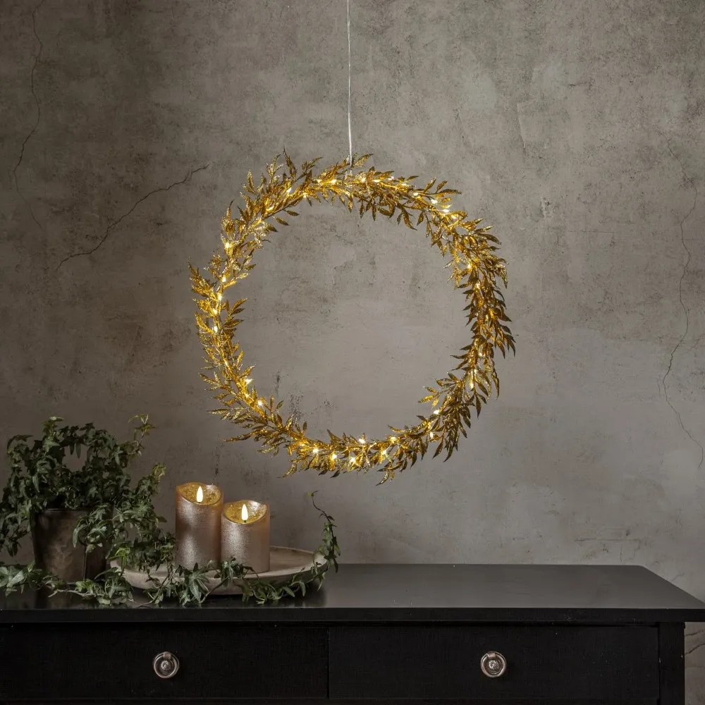 Złoty wianuszek dekoracyjny LED z drobnymi listkami i ciepłym światłem, zawieszony nad czarną komodą udekorowaną świecami i zielenią, na tle ściany o naturalnej fakturze.