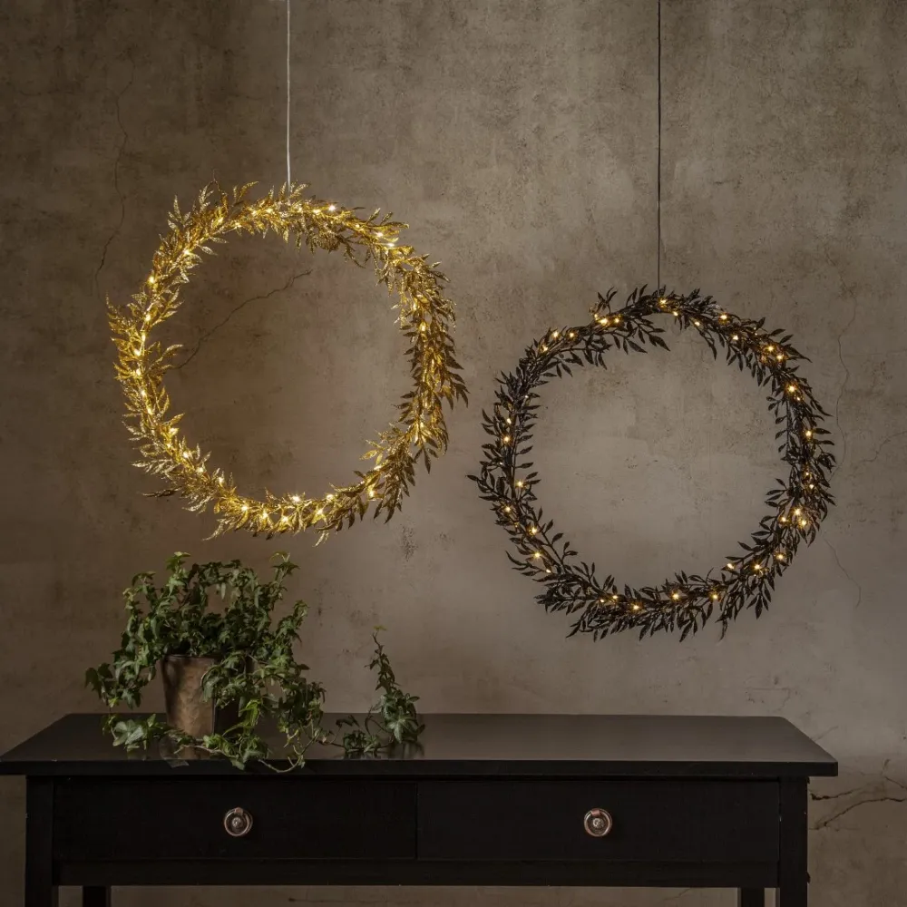 Złoty i czarny wianuszek dekoracyjny LED o listkowej formie i ciepłym świetle, zawieszone obok siebie na tle ściany z delikatną fakturą, nad komodą z zieloną rośliną.