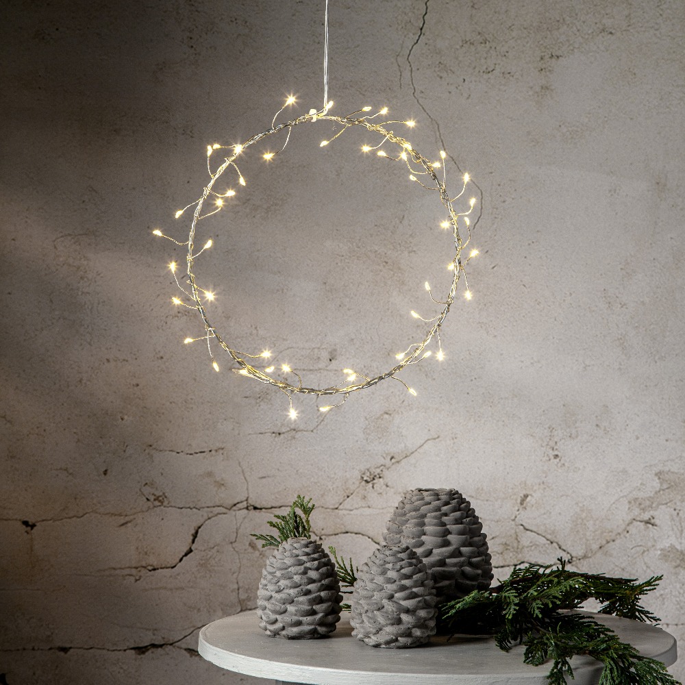 Wianek LED na subtelnej, metalowej ramie zawieszony na tle jasnej, lekko postarzonej ściany, roztaczający świąteczny blask nad stołem ozdobionym szyszkami i zielonymi gałązkami.