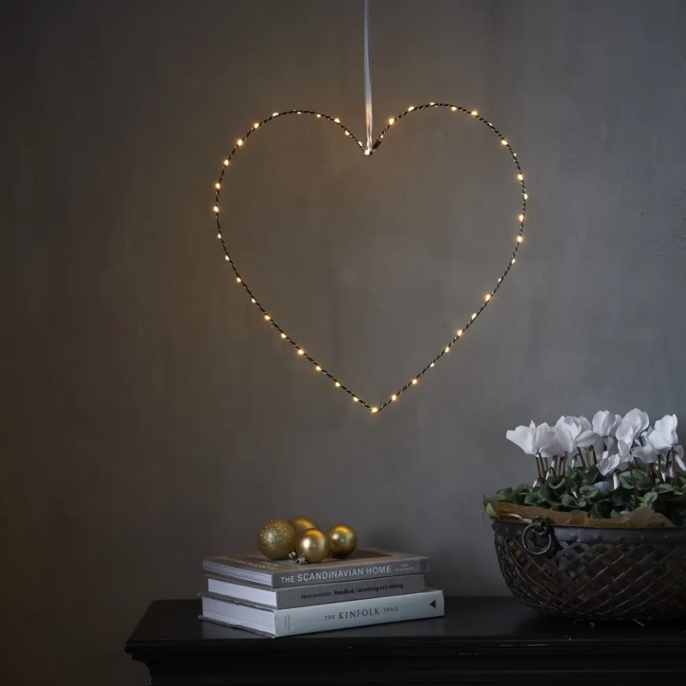 Delikatne serce LED zawieszone na jasnej wstążce, subtelnie rozświetlające wnętrze ciepłym blaskiem. Dekoracja tworzy przytulny nastrój nad stolikiem z książkami i złotymi akcentami.