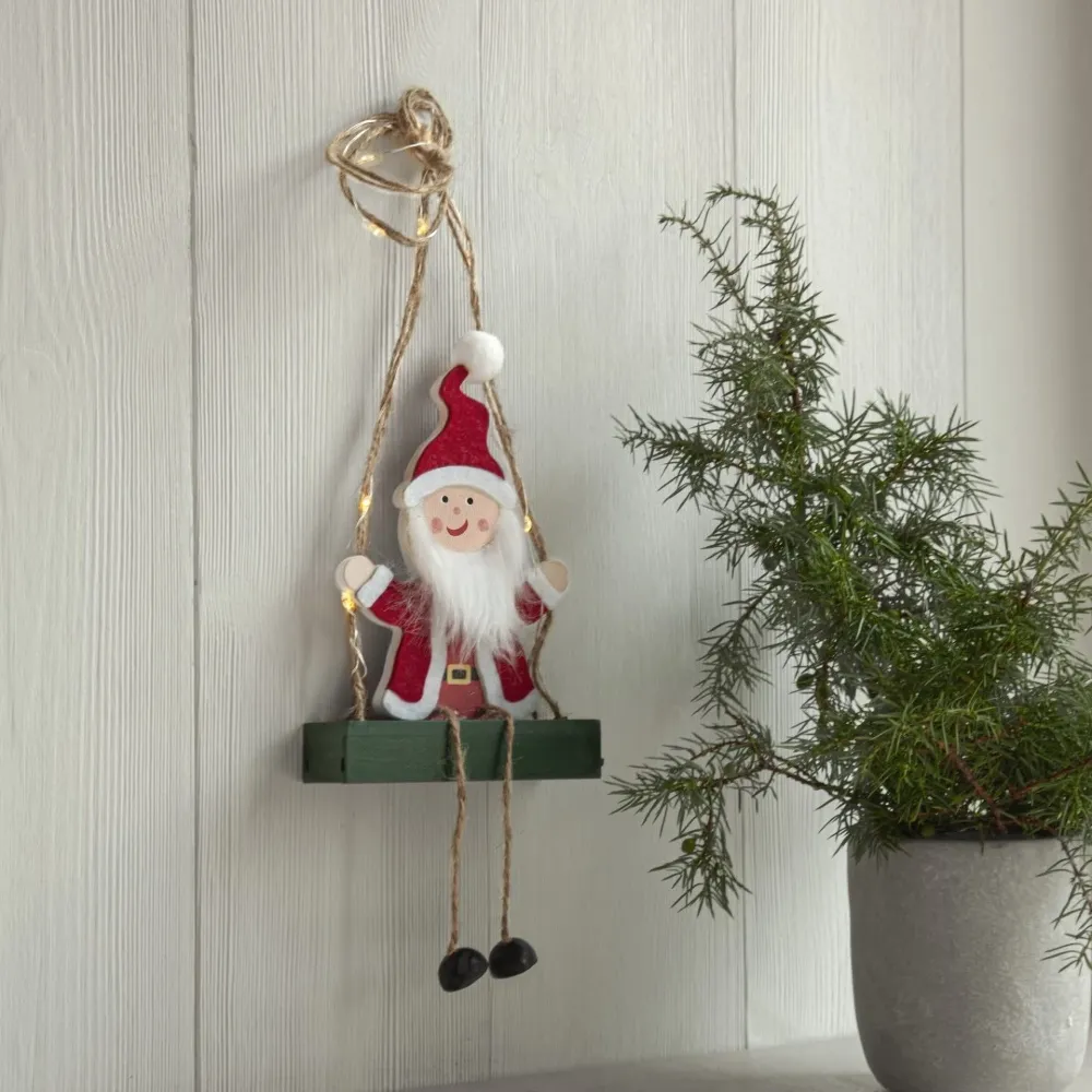 Drewniana dekoracja LED z Mikołajem siedzącym na huśtawce, zawieszona na naturalnym sznurku na jasnej, drewnianej ścianie; obok stoi zielona roślina w szarej donicy.