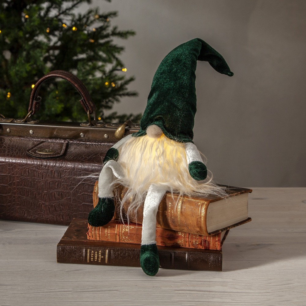 Zielony skrzat z długą czapką i podświetlaną brodą siedzący na stosie starych książek. Obok widać brązową walizkę i fragment świątecznego drzewka z delikatnymi światełkami.
