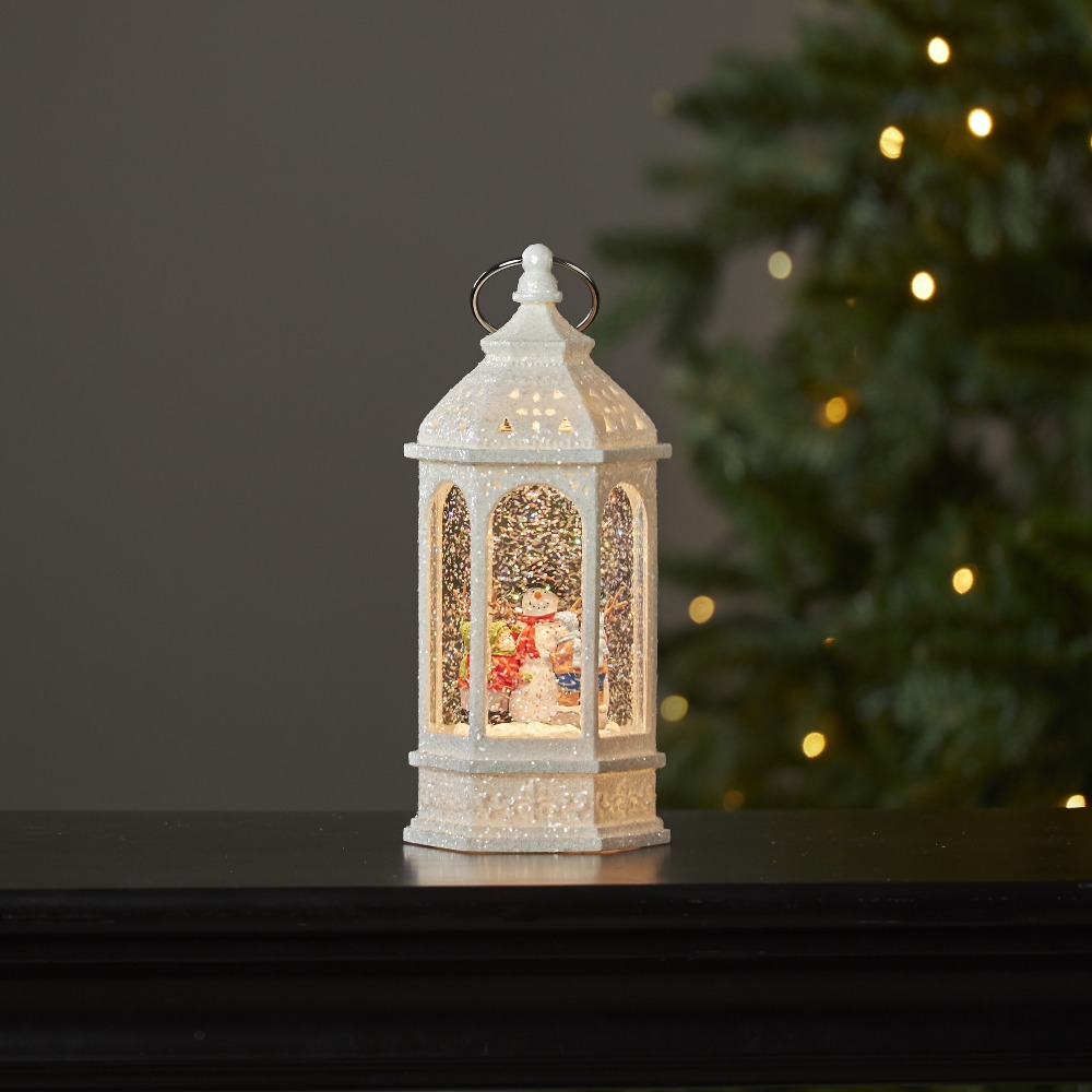 Podświetlana świąteczna latarenka wodna LED w białym, brokatowym wykończeniu, z bałwankiem i dziećmi w wirującym „śniegu”, ustawiona na ciemnym blacie na tle choinki z ciepłymi lampkami.