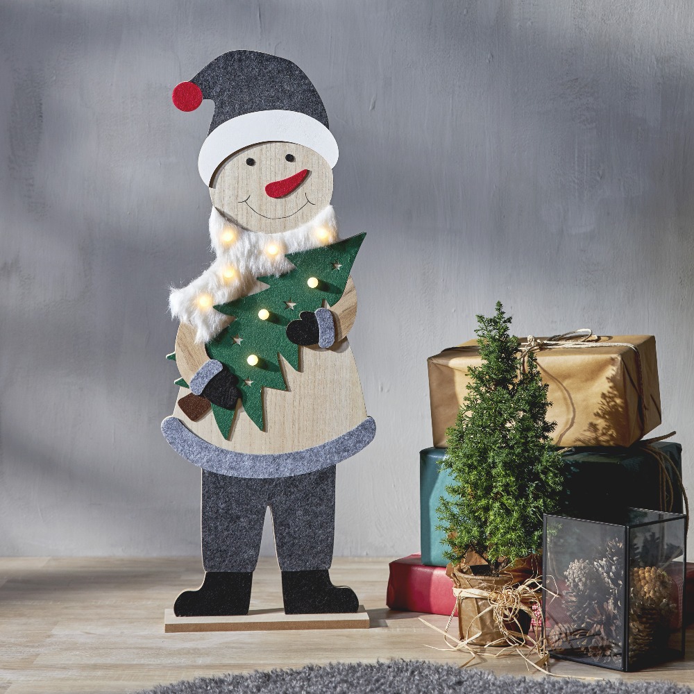 Drewniana dekoracja świąteczna LED – uśmiechnięty bałwanek z choinką, stojący obok prezentów i małej choinki w przytulnym, domowym wnętrzu, idealna ozdoba na święta.
