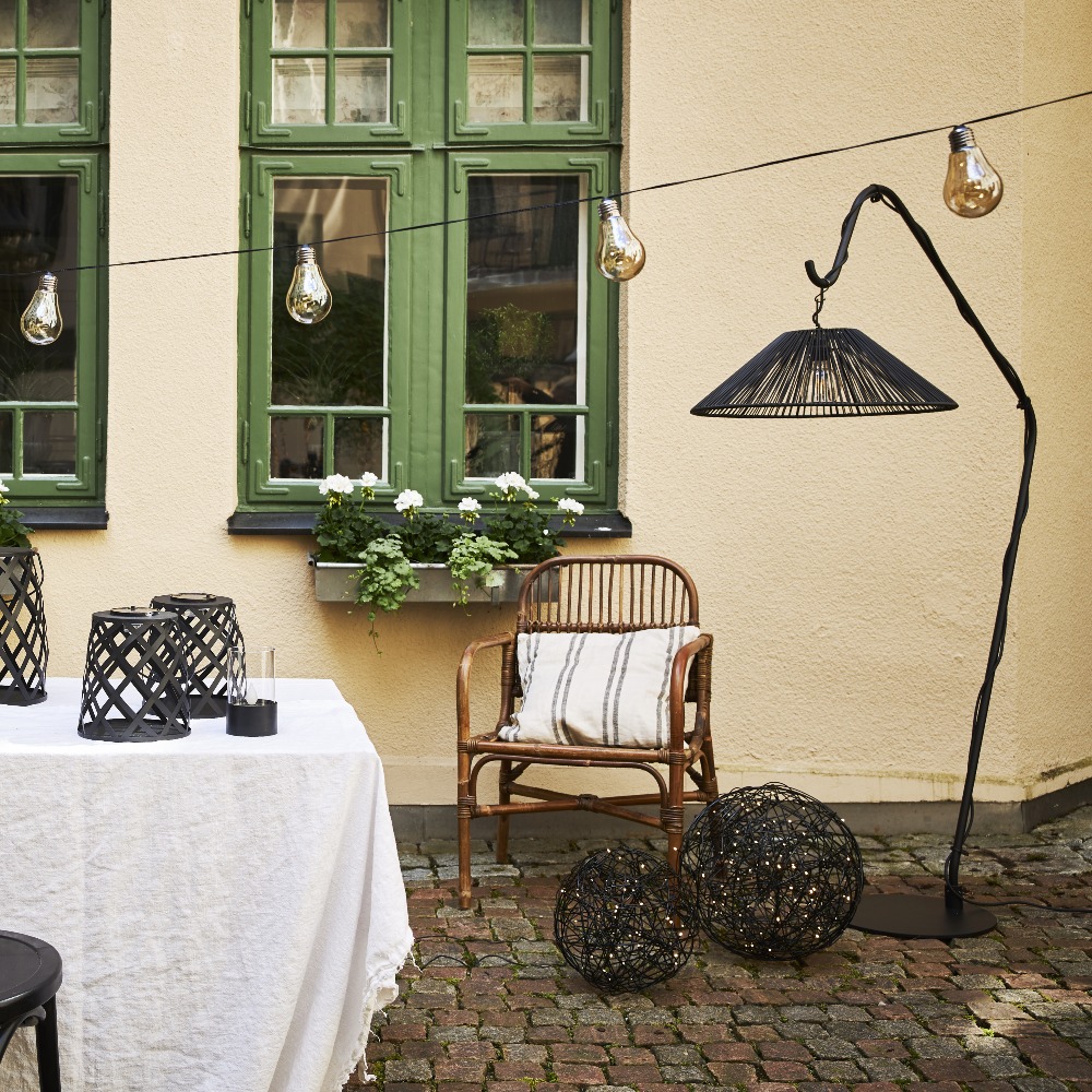 Dwie czarne kule z metalowego drutu ustawione na kamiennym podłożu na dziedzińcu przy ścianie budynku. Obok stoi lampa ogrodowa i meble, ażurowe formy dopełniają przytulną aranżację strefy wypoczynku.
