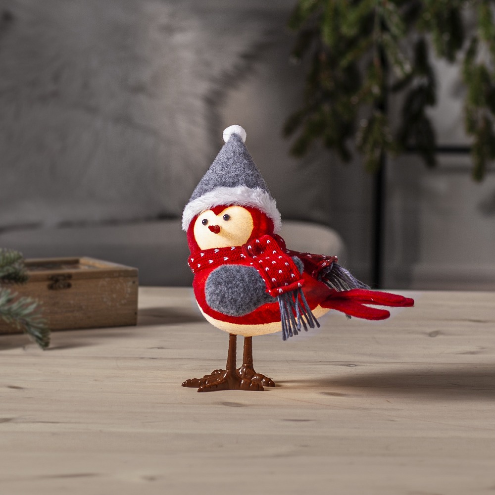 Świąteczny ptaszek dekoracyjny w czerwonym stroju z szalikiem i szarą czapką, stojący na jasnym drewnianym stole. W tle miękkie tkaniny i zimowe gałązki, tworzące ciepłą, domową aranżację.