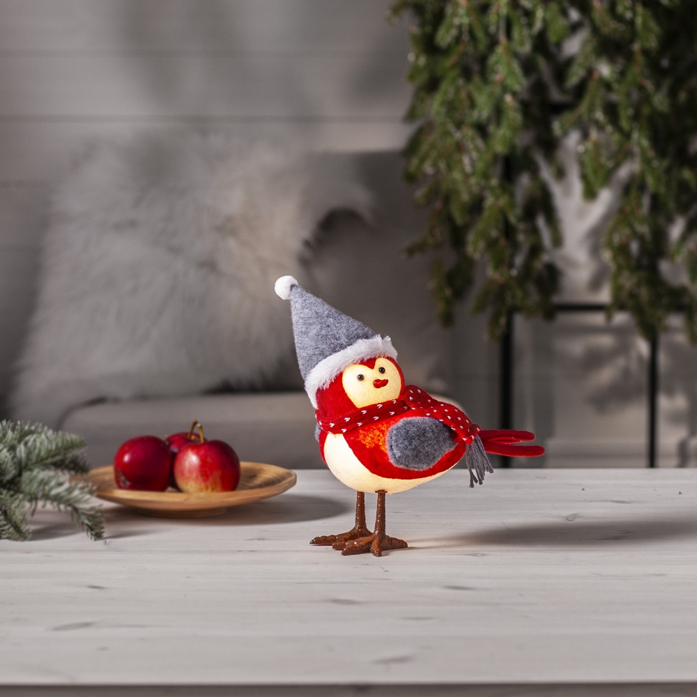 Ptaszek dekoracyjny w czerwonym stroju z szalikiem i szarą czapką, ustawiony na jasnym drewnianym stole. W tle zimowa aranżacja z gałązkami i owocami, tworząca ciepły, świąteczny klimat wnętrza.
