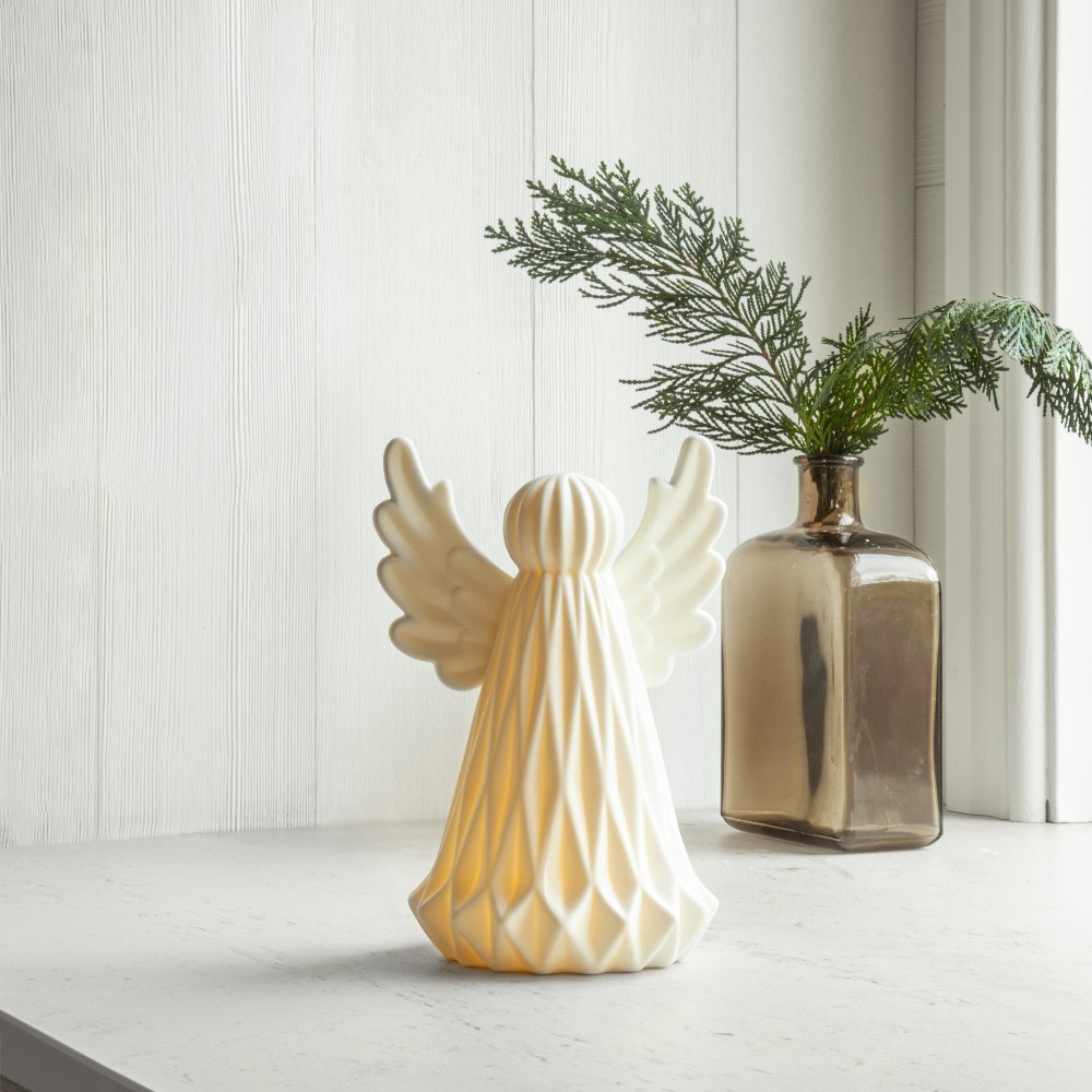 Biała ceramiczna dekoracja LED anioła, świecąca delikatnym światłem, ustawiona na jasnym parapecie obok szklanej butelki z zieloną gałązką; naturalne światło i spokojna, domowa atmosfera.