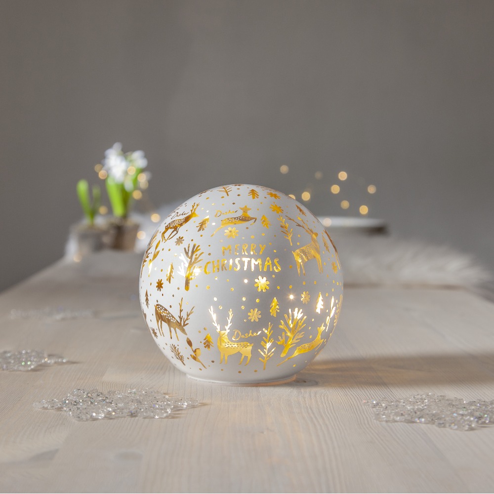 Szklana dekoracja stołowa LED w formie kuli z napisem „Merry Christmas” i złotymi motywami jeleni oraz drzew, świecąca ciepłym światłem, ustawiona na jasnym stole; delikatna, świąteczna atmosfera.