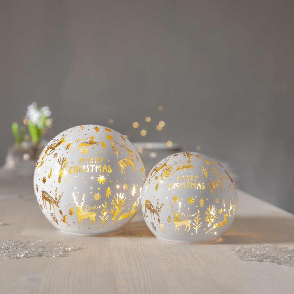 Dwie szklane dekoracje stołowe LED w kształcie kul z napisem „Merry Christmas” i złotymi motywami świątecznymi, świecące ciepłym światłem, ustawione na jasnym stole; spokojna, przytulna kompozycja.