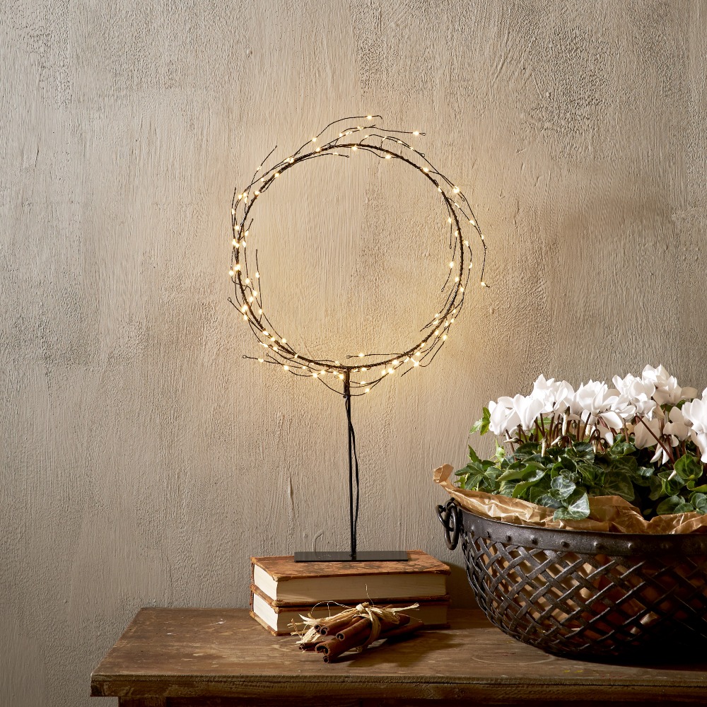 Dekoracyjny wieniec LED osadzony na smukłej podstawie, ustawiony na drewnianym meblu; ciepłe punkty światła, w tle książki i misa z białymi kwiatami. Spokojna, przytulna ozdoba do wnętrza.