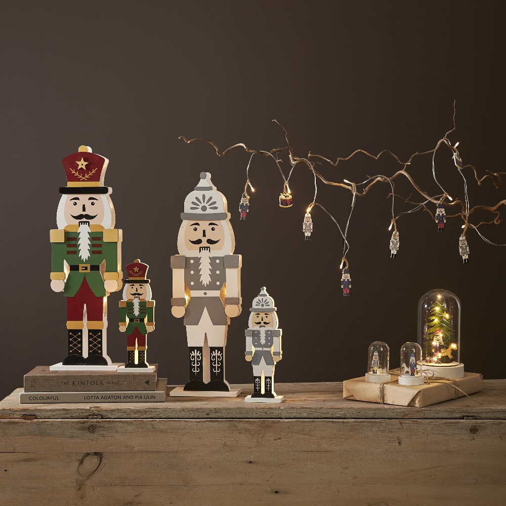Kilka drewnianych figurek dziadka do orzechów w różnych rozmiarach, ustawionych na stole wśród dekoracyjnych światełek. Subtelne podświetlenie tworzy spokojny, nastrojowy klimat wnętrza.