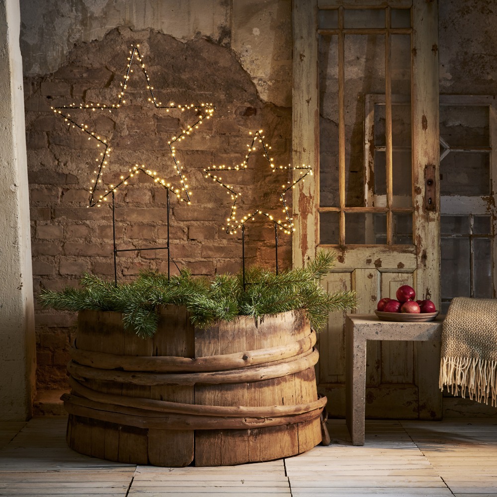 Dwie gwiazdy LED na metalowych stelażach z ciepłym światłem, ustawione w drewnianej beczce z zielonymi gałązkami. Przytulna świąteczna aranżacja w rustykalnym wnętrzu.