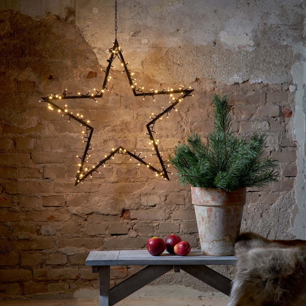 Wisząca gwiazda LED z drobnymi punktami świetlnymi zawieszona na łańcuchu; ciepłe światło tworzące przytulną aranżację na tle ceglanej ściany, obok donicy z zieloną rośliną i drewnianej ławki.
