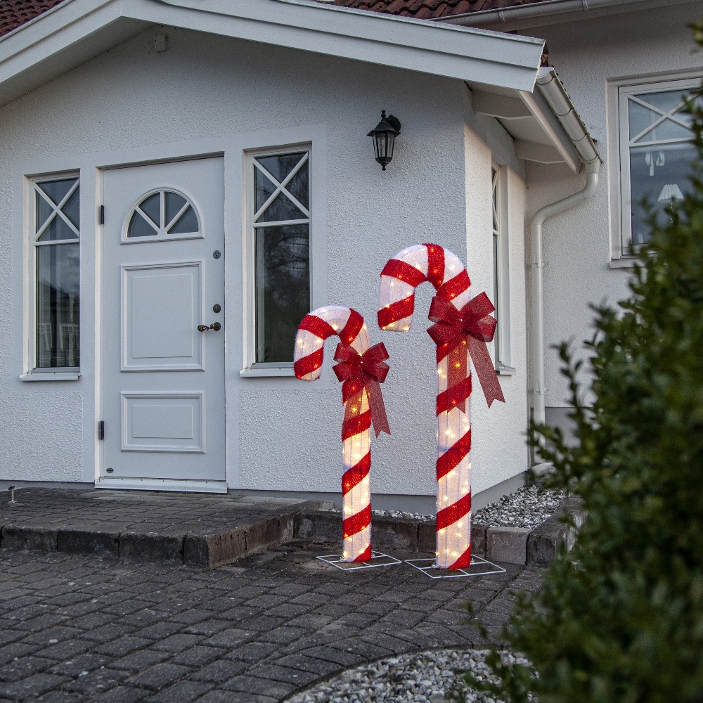 Dwie podświetlane laski cukrowe LED w czerwono-białe pasy z dekoracyjnymi czerwonymi kokardami, ustawione przed wejściem do domu na kostce brukowej; tworzą elegancką, spokojną dekorację świąteczną elewacji.