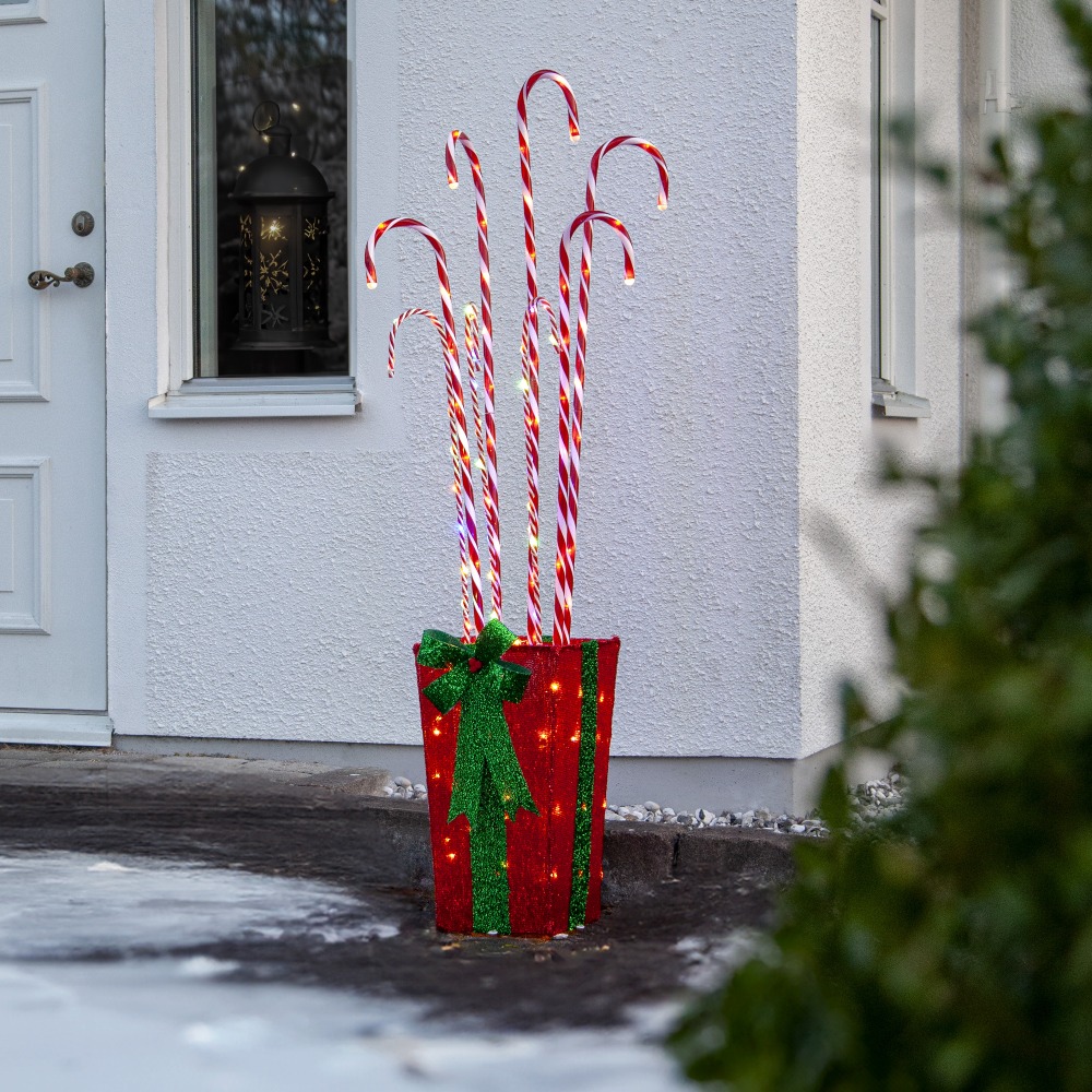 Świąteczna dekoracja LED w formie czerwonego pojemnika z zieloną kokardą, z którego wychodzą podświetlane czerwono-białe laski cukrowe, ustawiona przed wejściem do domu.