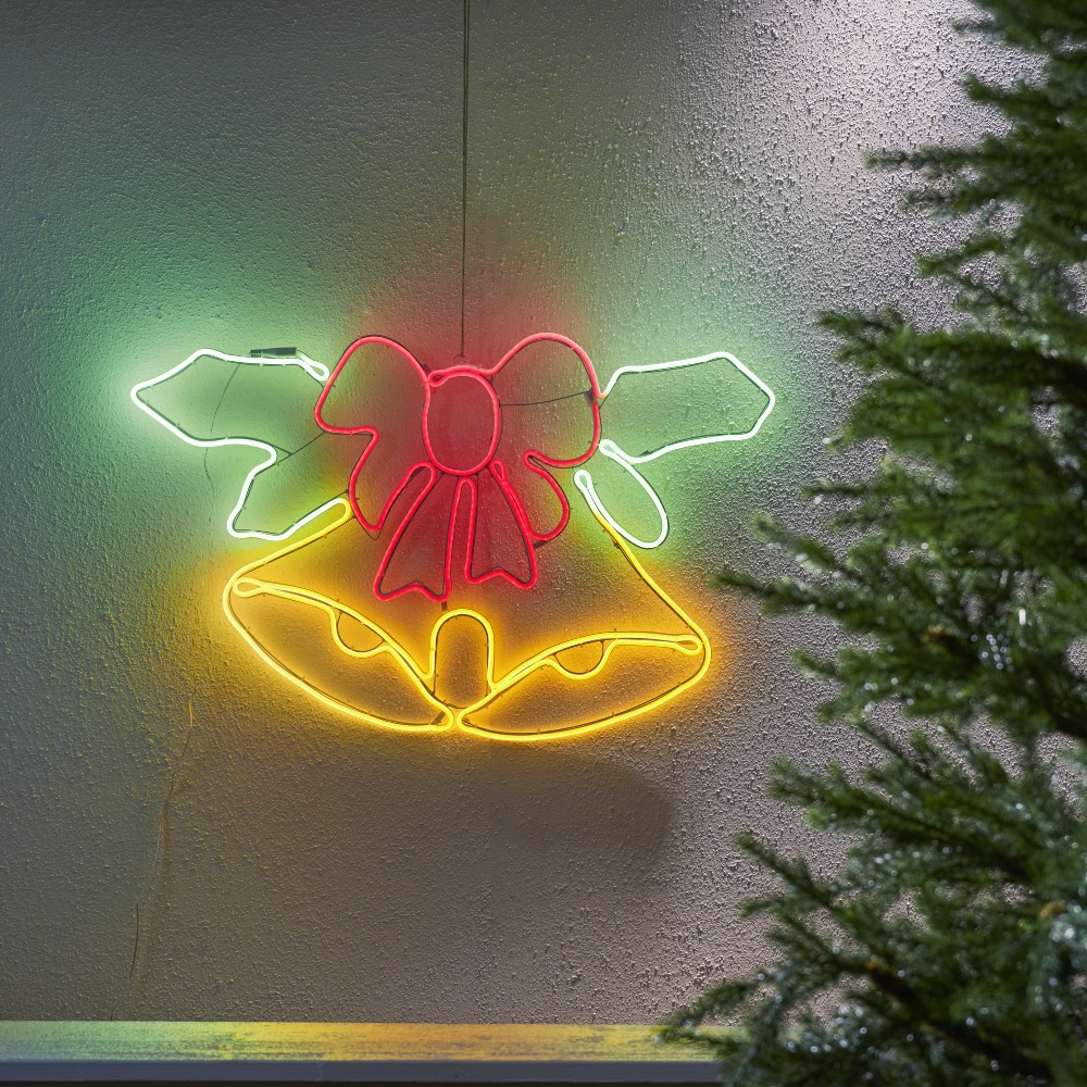 Dekoracja neon LED przedstawiająca dzwonki z kokardą, zawieszona na ścianie obok choinki. Ciepłe, kolorowe światło tworzy wyraźny kontur i harmonijny, świąteczny nastrój.