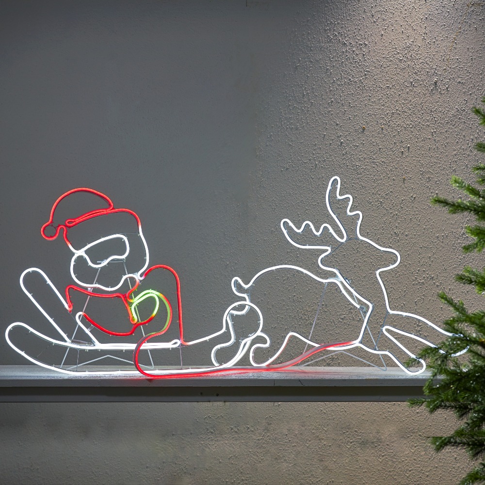 Dekoracja świetlna neon LED przedstawiająca Świętego Mikołaja na saniach z reniferem, ustawiona na półce przy ścianie. Białe, czerwone i zielone światło tworzy wyraźny kontur sceny o świątecznym charakterze.