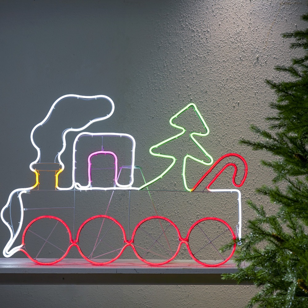 Dekoracja neon LED z motywem świątecznego pociągu, zamontowana przy ścianie obok choinki. Czerwone, zielone i białe światło podkreśla kształt lokomotywy i nadaje całości ciepły, świąteczny charakter.