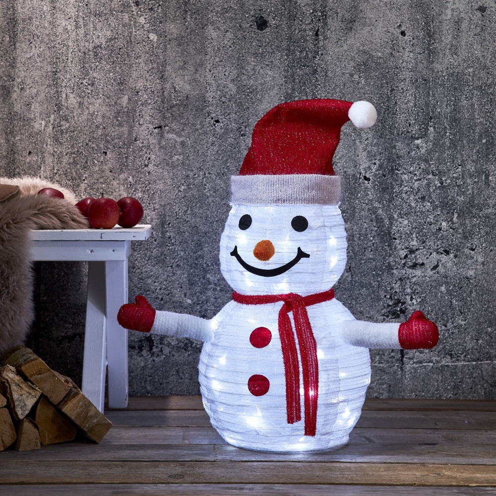 Dekoracja świetlna LED w kształcie bałwanka z czerwoną czapką i szalikiem, ustawiona we wnętrzu przy ścianie. Delikatne białe światło podkreśla strukturę postaci i tworzy przytulny, świąteczny klimat.