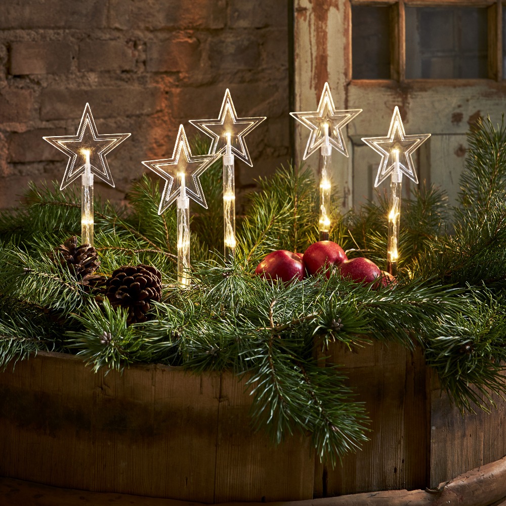 Świąteczna aranżacja z pięcioma dekoracjami LED w kształcie gwiazd na palikach ustawionymi wśród iglastych gałązek i ozdób delikatne światło buduje spójny i przytulny efekt wizualny.