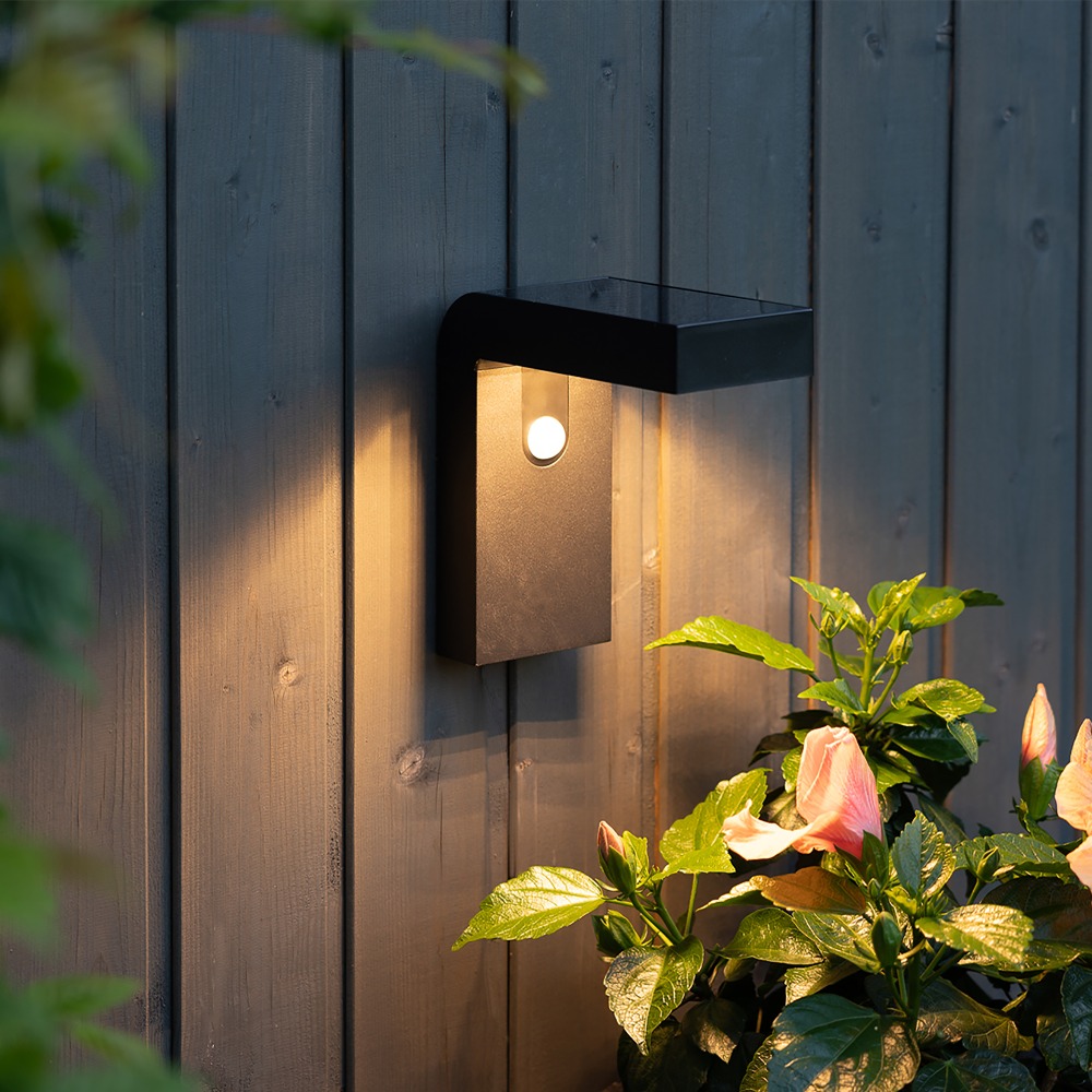 Czarny solarny kinkiet ogrodowy LED VICI z czujnikiem ruchu PIR zamontowany na drewnianej elewacji ciepłe światło oświetlające rośliny przy ścianie.