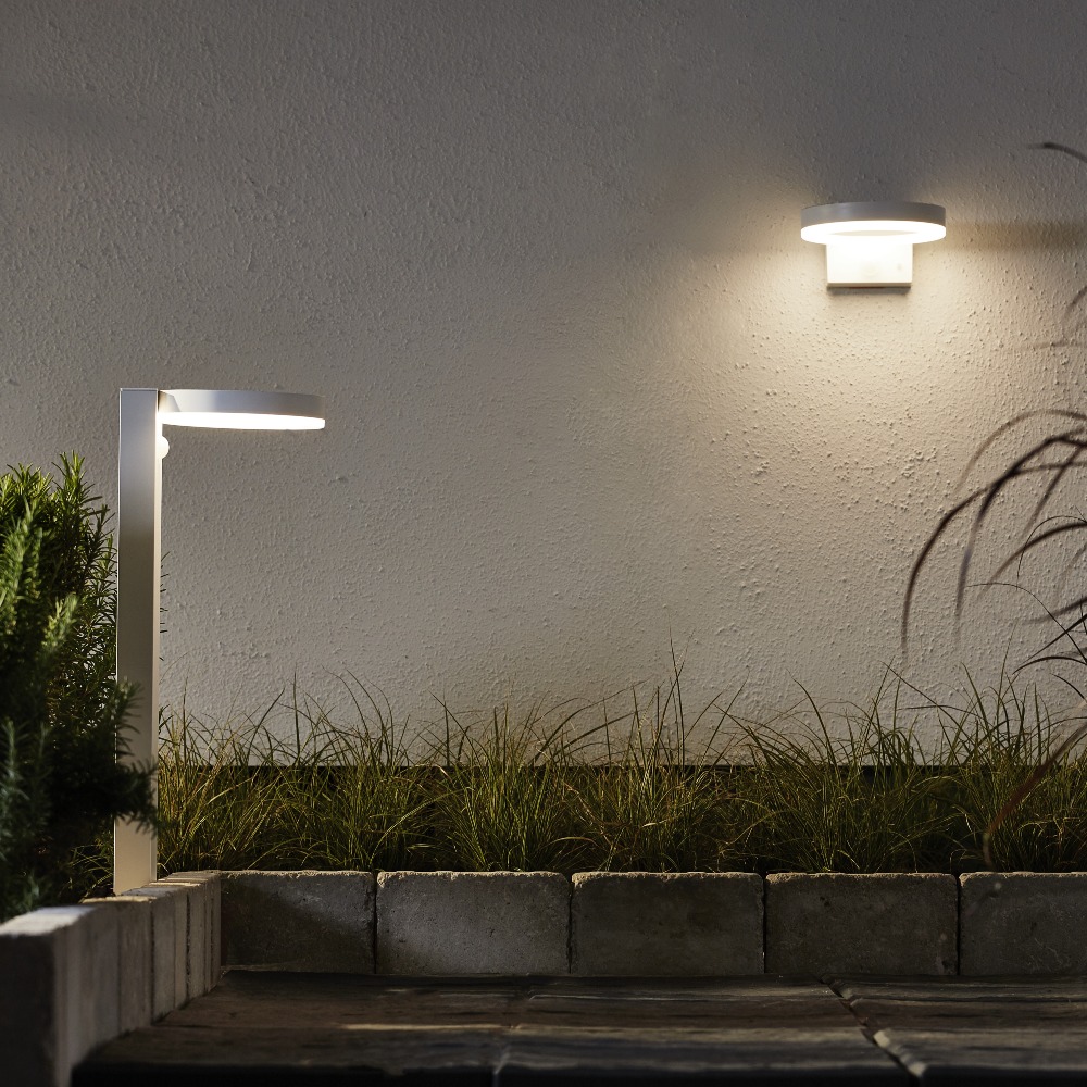 Solarna lampa ogrodowa LED słupek oraz kinkiet ścienny oświetlające ścieżkę i elewację budynku ciepłe światło w aranżacji ogrodu nocą.