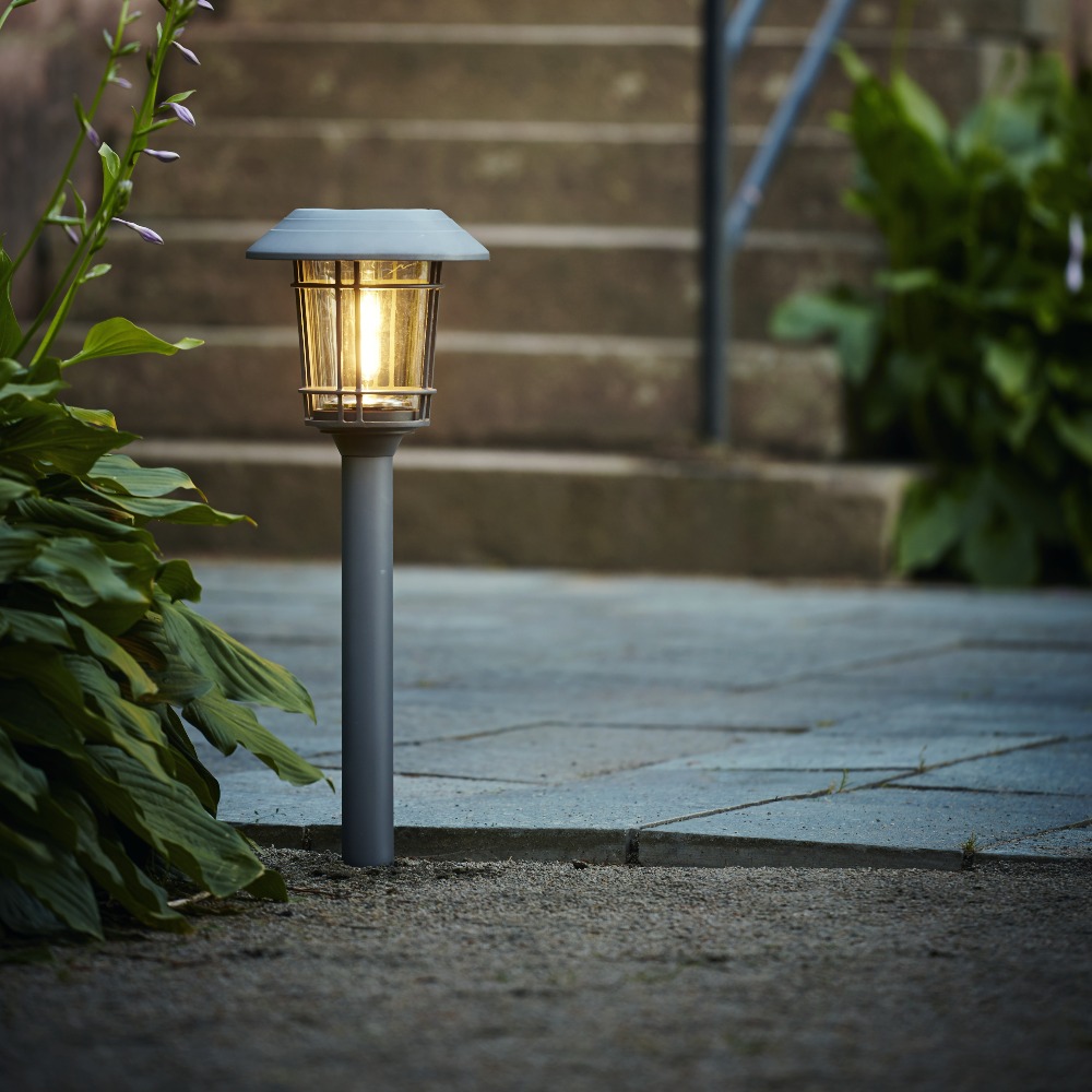 Lampa solarna ogrodowa w formie słupka z daszkiem i przeszkloną osłoną, emitująca ciepłe światło. Ustawiona w ziemi przy kamiennych schodach i roślinach, delikatnie rozświetla przestrzeń po zmroku.