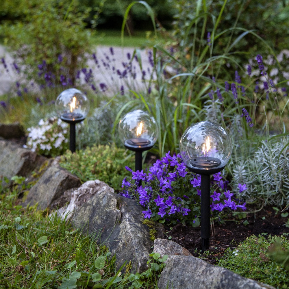 Solarne lampy ogrodowe LED z przezroczystym kulistym kloszem wbijane w ziemię, zamontowane w rabacie wśród kamieni i fioletowych kwiatów, dekoracyjne oświetlenie ogrodu i ścieżki.