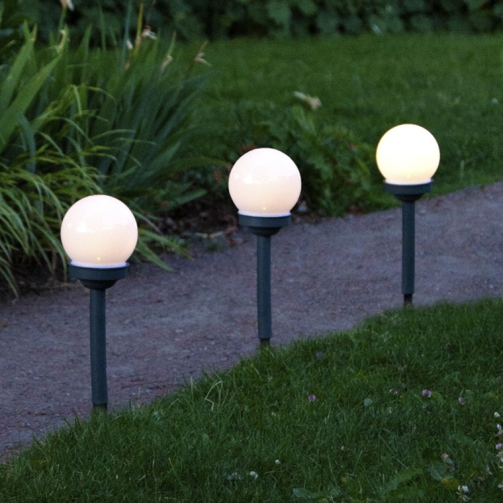 Solarne lampy ogrodowe z kulistym mlecznym kloszem ustawione wzdłuż ścieżki w ogrodzie, delikatnie oświetlające trawnik po zmroku.