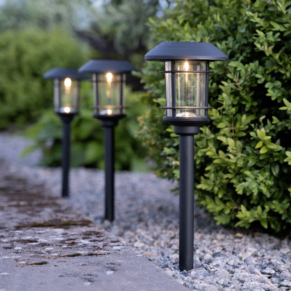 Trzy czarne solarne lampy ogrodowe z przezroczystymi kloszami osłoniętymi kratką, ustawione przy betonowej ścieżce obok pasa żwiru i zielonego żywopłotu.