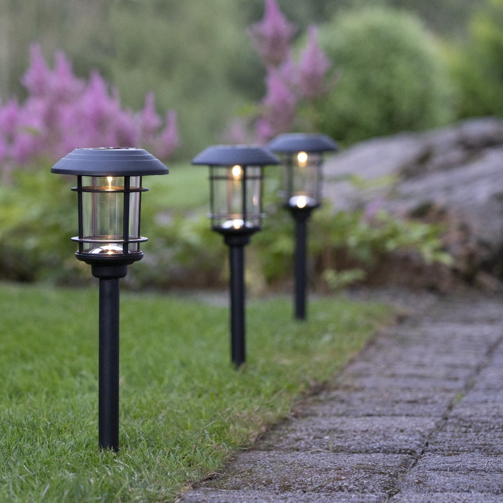 Trzy czarne solarne lampy ogrodowe z przezroczystymi kloszami osłoniętymi kratką, ustawione w trawie wzdłuż ścieżki z kostki brukowej, na tle roślin i dużego kamienia.