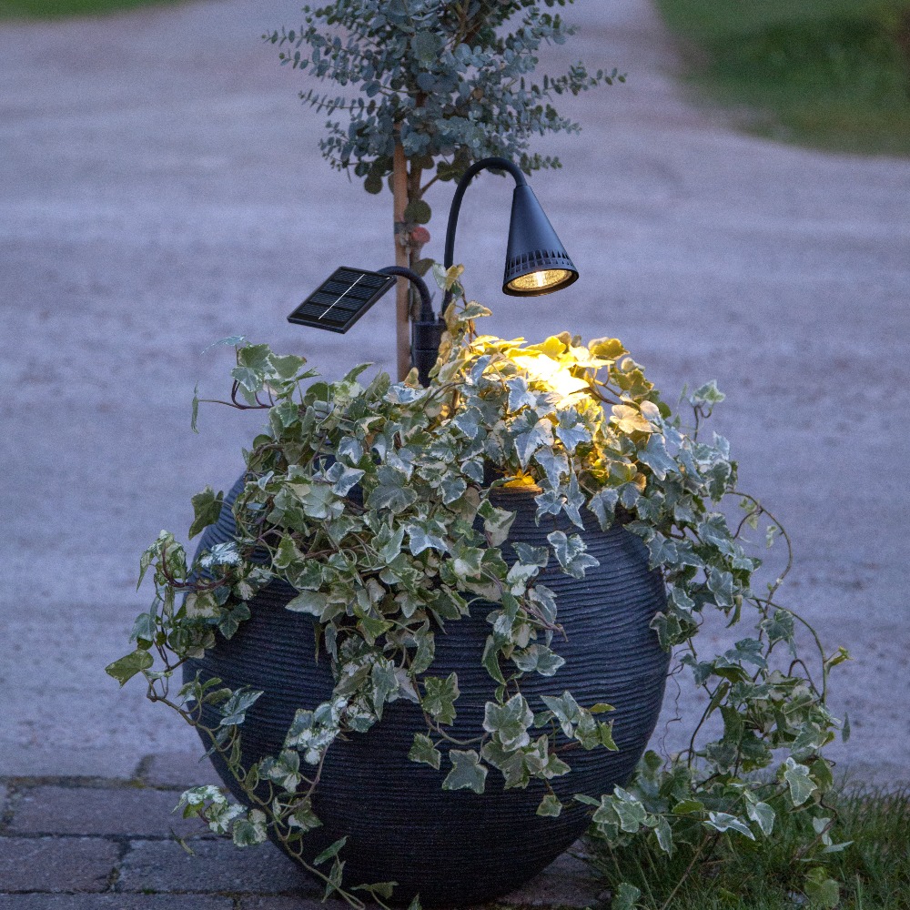 Solarny reflektor ogrodowy LED zamontowany w donicy z roślinami, z głowicą świetlną i panelem solarnym osadzonymi na giętkich ramionach, emitujący ciepłe białe światło podkreślające zieleń.