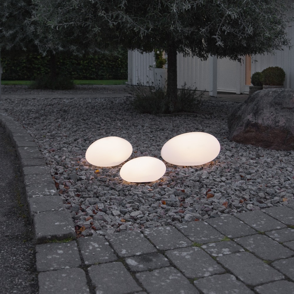 Solarne lampy ogrodowe LED w kształcie kamieni świecące ciepłym światłem ustawione na żwirowej rabacie przy ścieżce ogrodowej. Dekoracyjne oświetlenie solarne do ogrodu, na podjazd, rabaty i przy domu.
