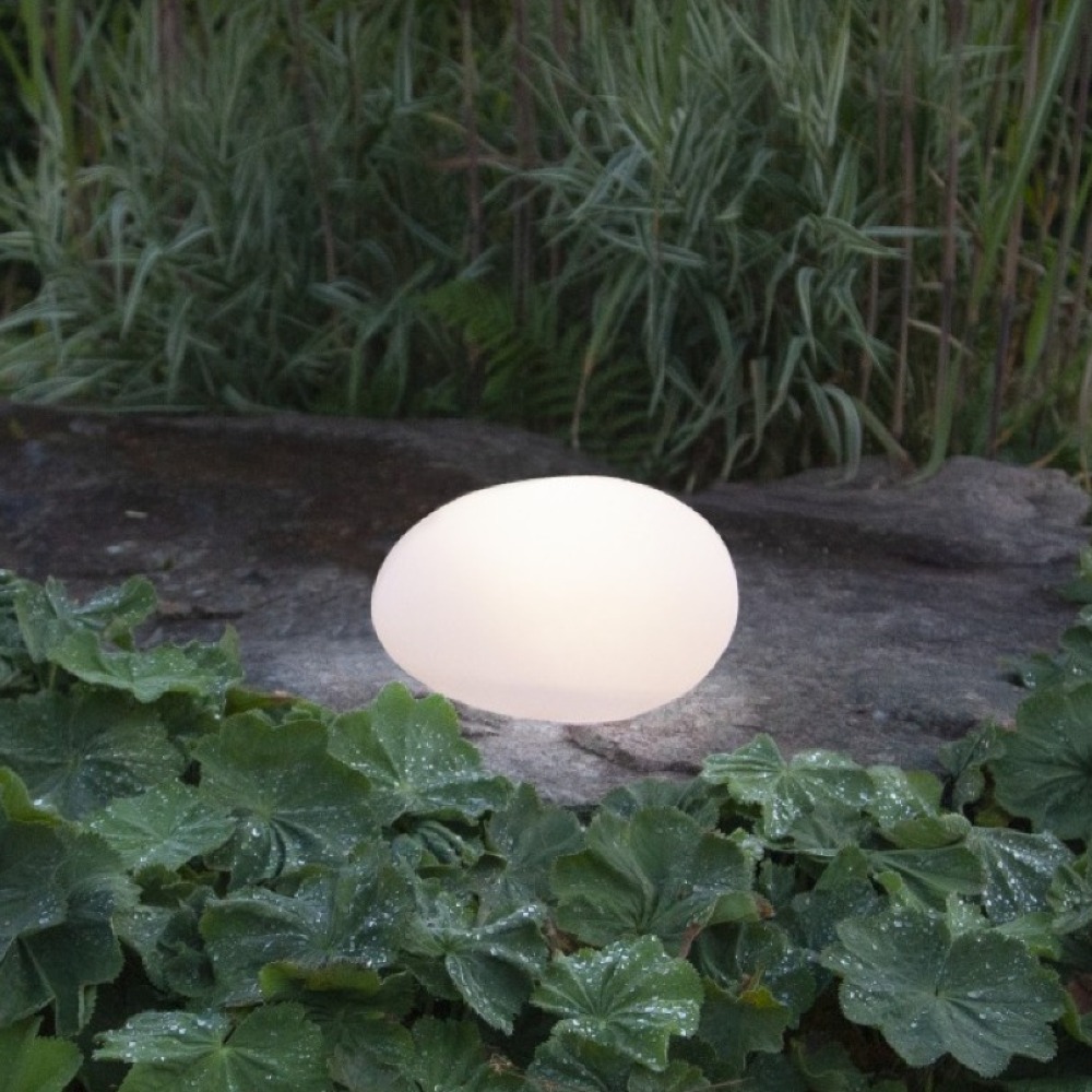 Lampa solarna LED w kształcie kamienia świecąca ciepłym światłem ustawiona na kamiennej płycie wśród roślin ogrodowych. Dekoracyjne oświetlenie solarne do ogrodu, na rabaty, ścieżki i taras.