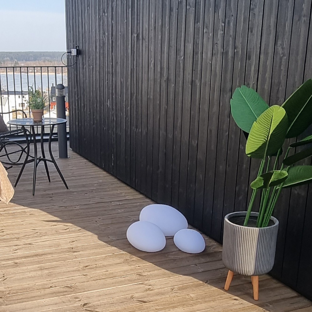 Solarne dekoracje LED w kształcie kamieni ustawione na drewnianym tarasie przy ścianie domu obok dużej donicy z rośliną. Dekoracyjne oświetlenie solarne do ogrodu, na taras, balkon lub patio.