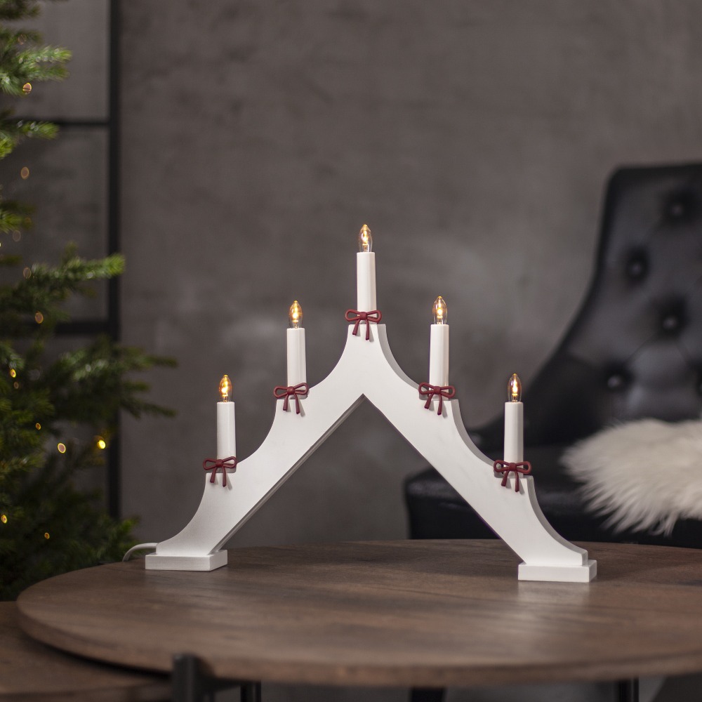 Biały świecznik dekoracyjny z pięcioma świecami ozdobionymi czerwonymi kokardkami, ustawiony na stole w salonie obok choinki.
