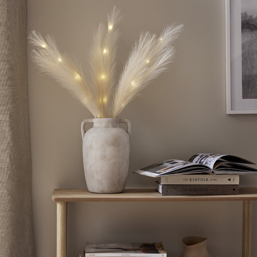 Pióra dekoracyjne LED w kolorze ecru z ciepłym światłem, ustawione w wazonie na półce obok książek w minimalistycznym wnętrzu.