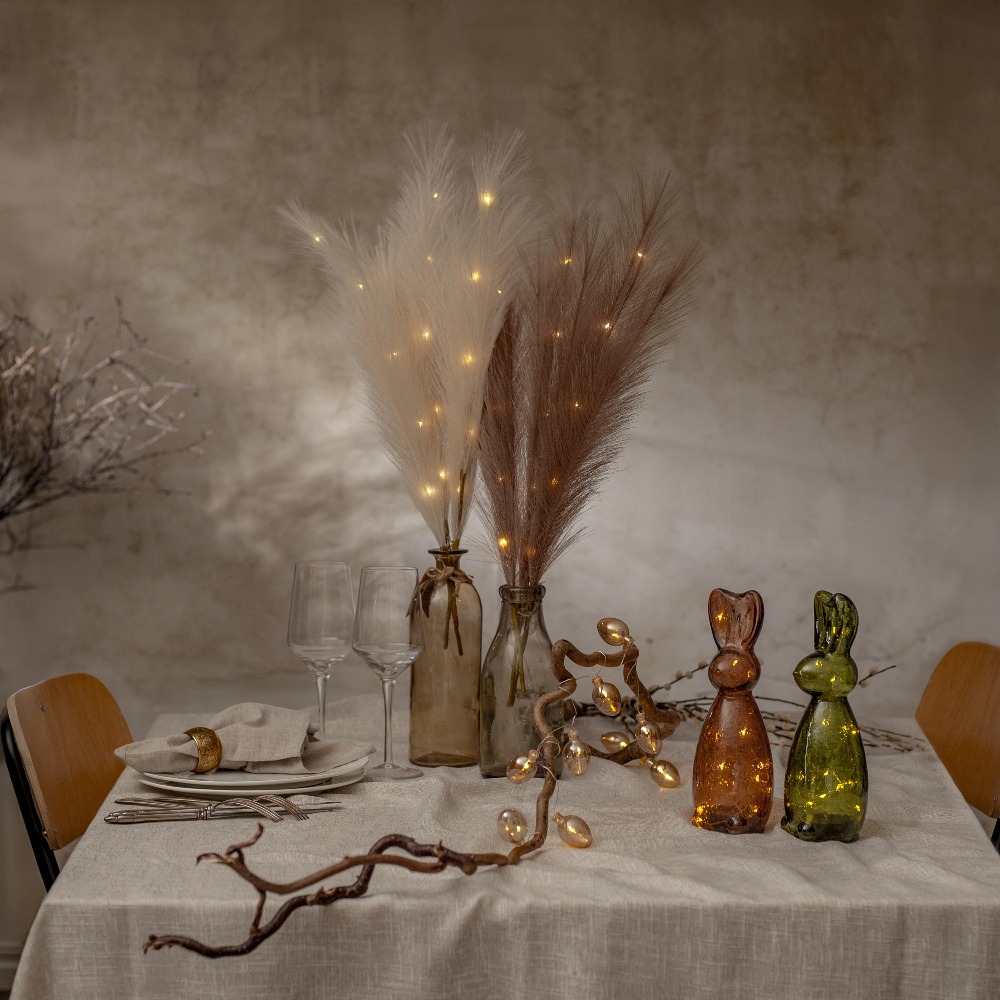 Pióra dekoracyjne LED w kolorze ecru i brązowym z ciepłym światłem, ustawione w szklanych wazonach na stole w eleganckiej aranżacji.