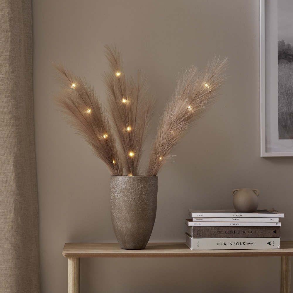 Brązowe pióra dekoracyjne z oświetleniem LED umieszczone w wazonie na półce obok książek stanowią klimatyczną dekorację z ciepłym światłem.