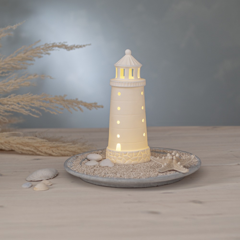 Biała porcelanowa latarnia morska LED na podstawie z piaskiem i muszlami ustawiona na stole jako element aranżacji wnętrza emitująca ciepłe światło.