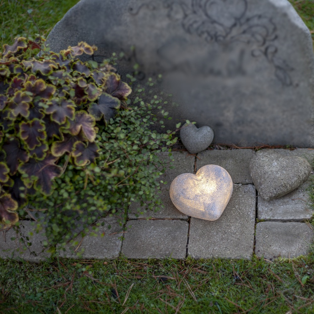 Dekoracyjne serce LED z efektem kamienia emitujące ciepłe światło, ustawione na nagrobku wśród roślin i kamieni, jako subtelna ozdoba pamięci.