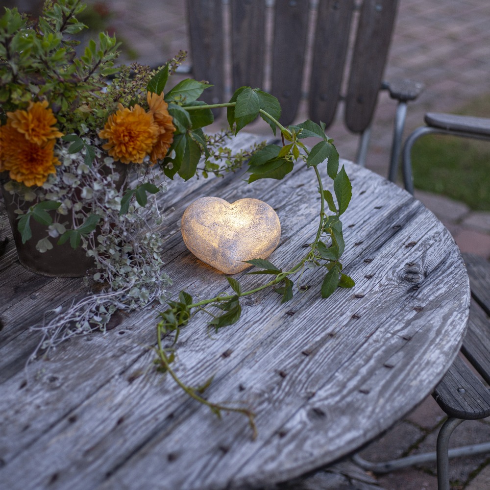 Dekoracyjne serce LED z efektem kamienia emitujące ciepłe światło, ustawione na drewnianym stole w ogrodzie, otoczone kwiatami i zielonymi pędami, jako nastrojowa ozdoba przestrzeni zewnętrznej.