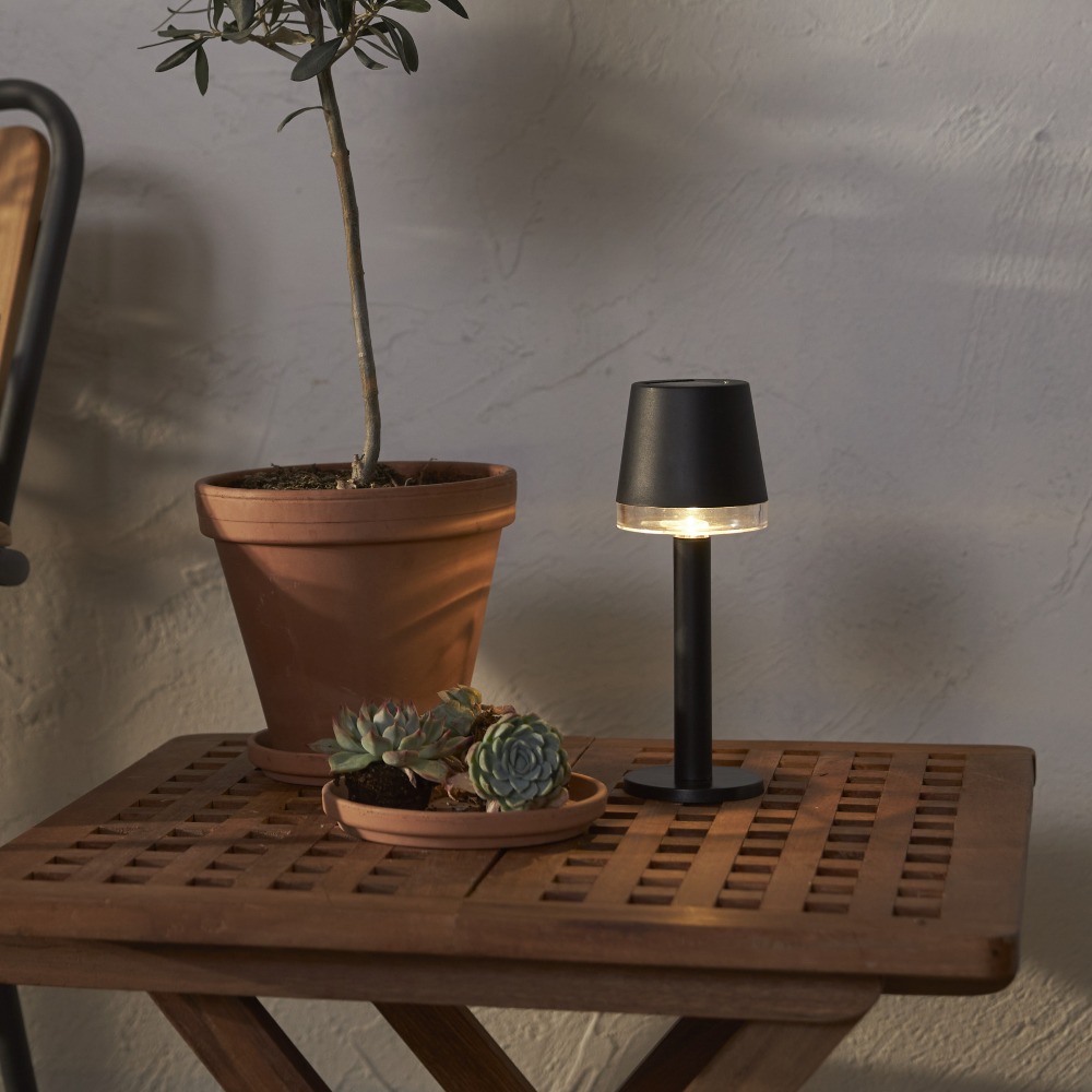 Solarna lampa stołowa LED z ciepłym światłem ustawiona na drewnianym stoliku obok roślin.