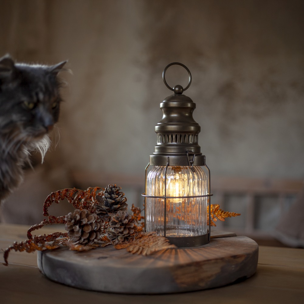 Stylowa lampa w kształcie latarni z ciepłym światłem, ustawiona na stole w towarzystwie naturalnych dekoracji, idealna do stworzenia nastrojowej aranżacji wnętrza.
