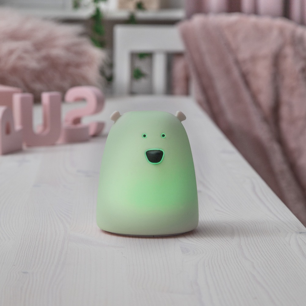 Lampka nocna w kształcie misia emitująca zielone światło, ustawiona na stoliku jako nastrojowe oświetlenie do pokoju dziecięcego.