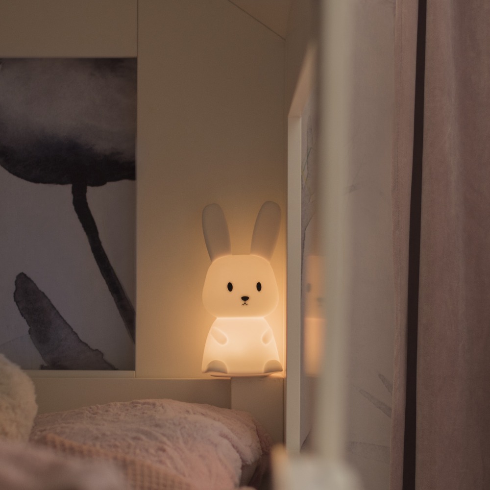 Lampka nocna króliczek świecąca ciepłym światłem, ustawiona przy łóżku jako spokojne oświetlenie nocne dla dziecka.